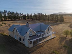 Aerial view of a charming country house with a blue roof and wraparound veranda surrounded by open grassland and tall pine trees.