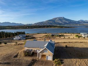 Aerial view of a gray-roofed lakeside house with a garage, surrounded by dry grassland, with a calm lake and mountains in the background.