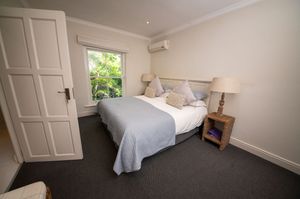 Bright bedroom with a double bed, light blue throw, white pillows, wooden nightstand with lamp, wall air conditioner and a garden window.