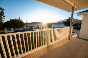 Sunset view from a spacious covered balcony with white railings overlooking neighboring houses, green trees and distant mountains.