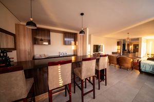 Open-plan apartment interior with a dark wood breakfast bar, cushioned red-framed stools, pendant lights and a lounge area beyond.