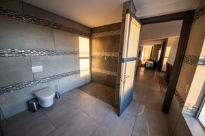 Modern bathroom with gray stone-look tiles, mosaic accent stripes, wall-mounted toilet, walk-in shower and open door to the adjoining room.