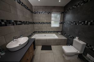 Modern bathroom with gray tile walls, mosaic accent stripes, a built-in bathtub, vessel sink with wooden vanity, and a white toilet.
