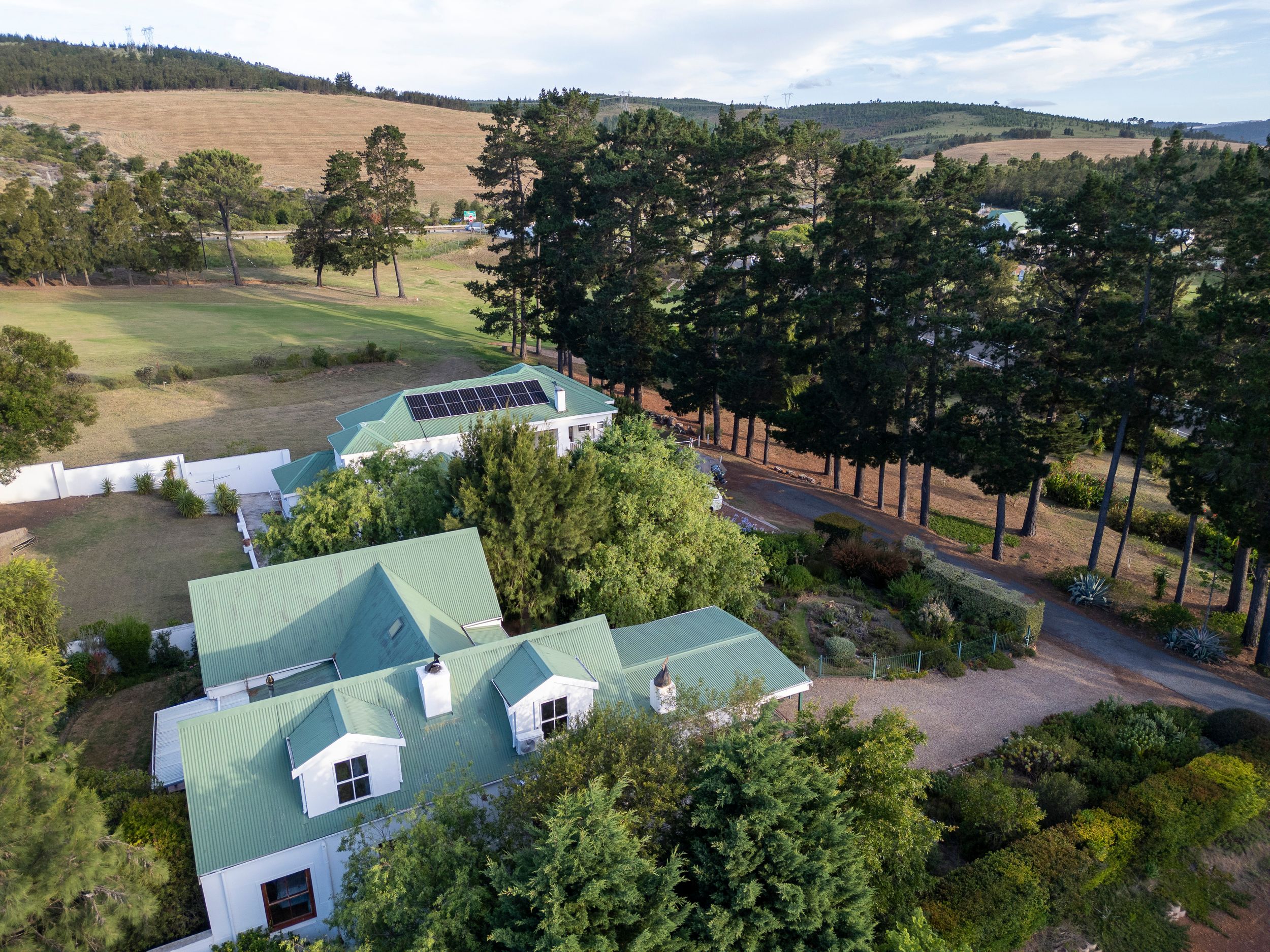 Aerial view of a white country house with green metal roofs, tall pine trees, manicured gardens and a gravel driveway amid rolling hills.