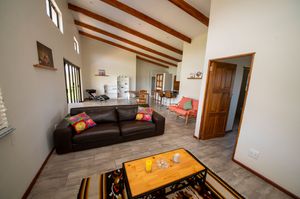 Spacious open-plan living room with vaulted beam ceiling, dark leather sofa with colorful pillows, wooden coffee table and cozy dining area.