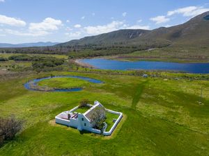 Aerial view of a whitewashed farmhouse with courtyard walls on green meadows beside a blue lake, with mountains rising in the background.