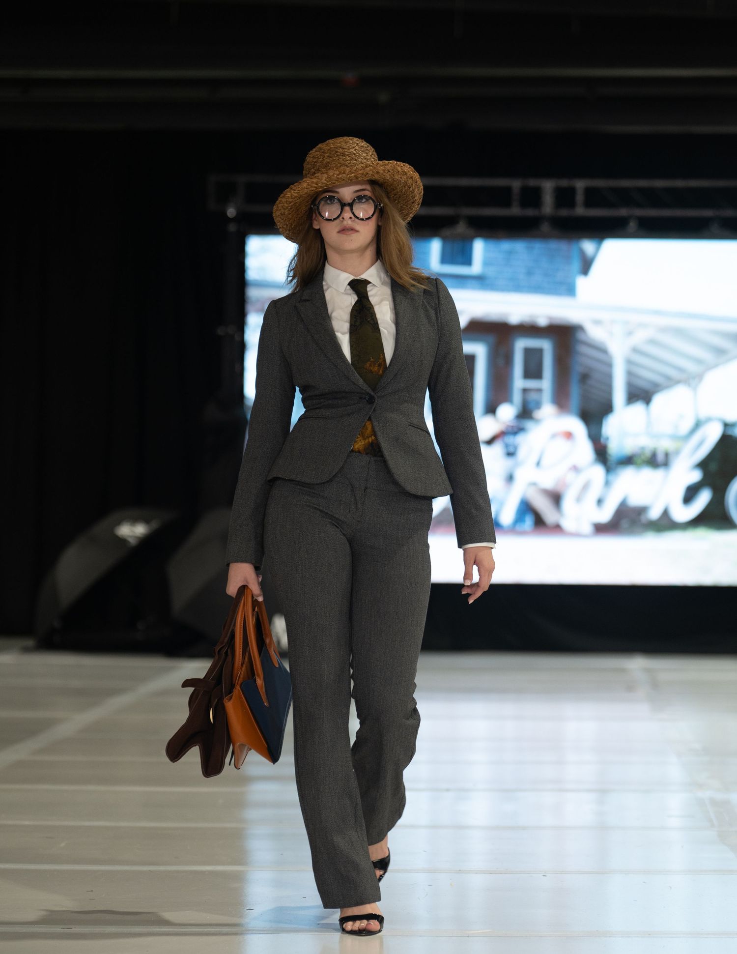 A model walks a fashion show runway in a gray tailored pantsuit, tie, round glasses and straw hat, carrying colorful leather bags.