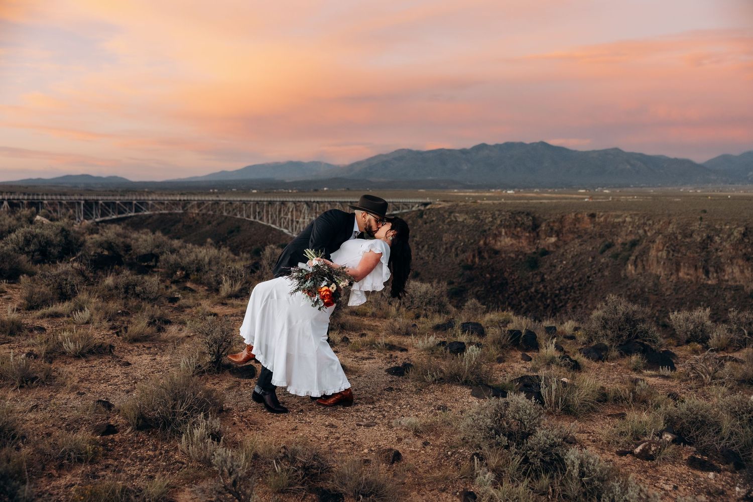 Couple sharing a romantic dip kiss at sunset with mountains and bridge backdrop in a desert landscape.