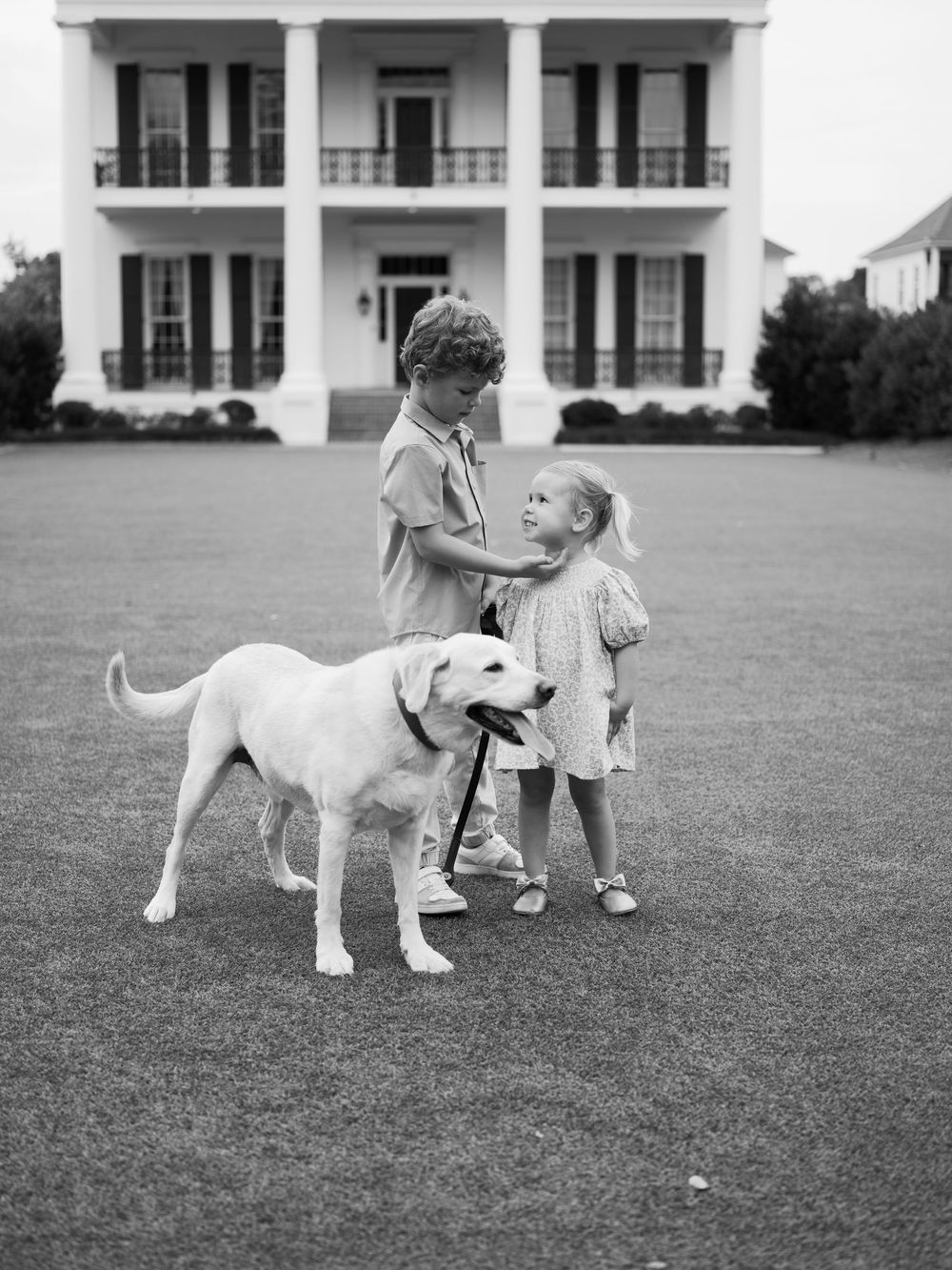 Black and white photo of two children with a yellow Labrador on lawn before a Southern plantation home.