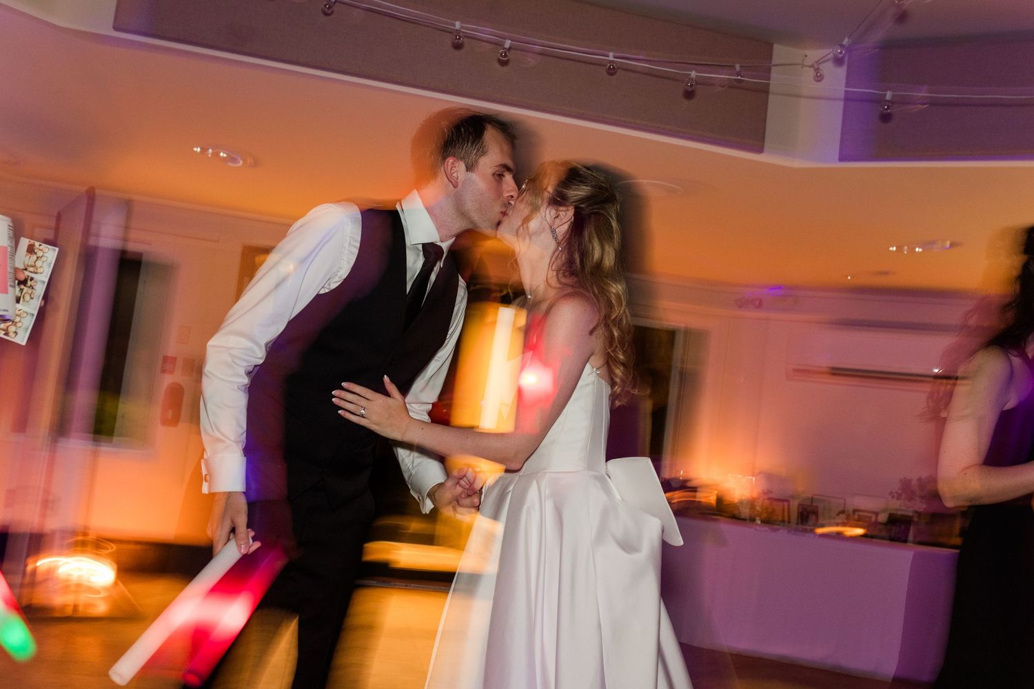 Bride and groom share a romantic first dance kiss at their wedding reception with colorful lights glowing around them.