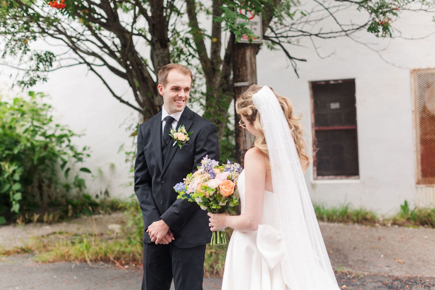Bride in white gown holding colorful bouquet faces smiling groom in black suit during outdoor first look wedding moment.