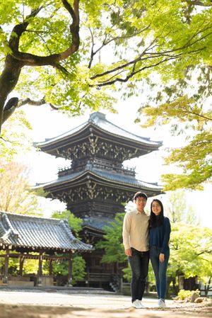 Smiling couple poses together beneath bright green maple leaves in front of a traditional Japanese temple pagoda on a sunny day.