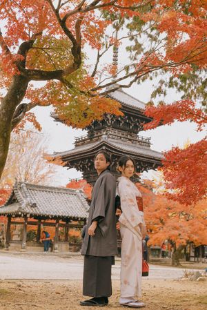 Two people in traditional Japanese kimono stand back to back before a historic pagoda surrounded by vibrant red autumn maple leaves.