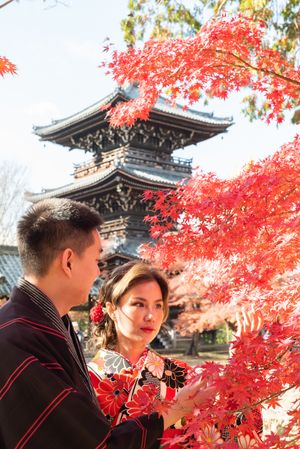 Couple in traditional kimono admire vibrant red maple leaves in front of a historic multi-tiered Japanese pagoda on a sunny autumn day.