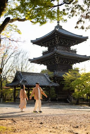 Couple in beige outfits walking hand in hand past a traditional Japanese three-story pagoda and temple hall surrounded by spring trees.