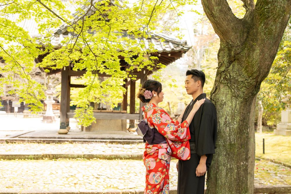 Couple in traditional Japanese kimono and yukata pose beside a large tree near a wooden temple pavilion surrounded by green autumn foliage.
