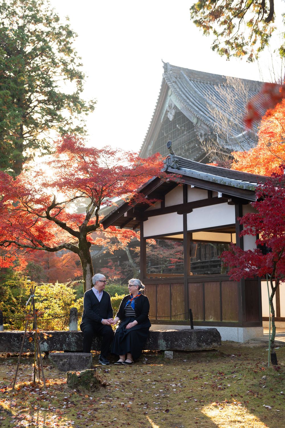 Two people sit and talk on a stone ledge beside a traditional Japanese temple building framed by vivid red autumn maple leaves.