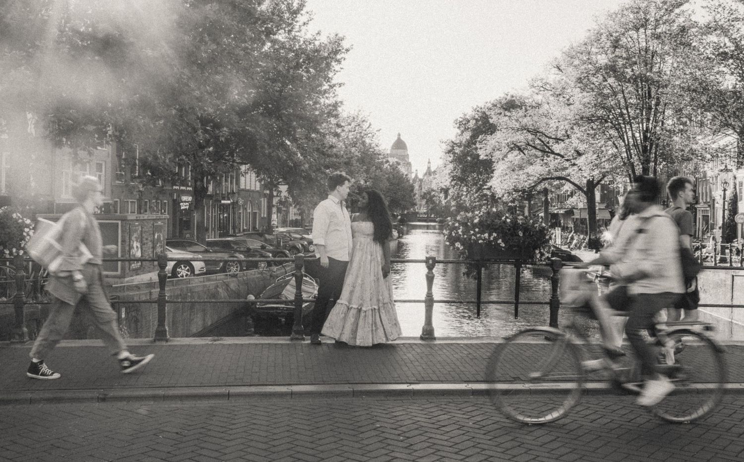 Black and white photo of a couple embracing on a canal bridge in Amsterdam as a cyclist and pedestrians pass by on a misty day.