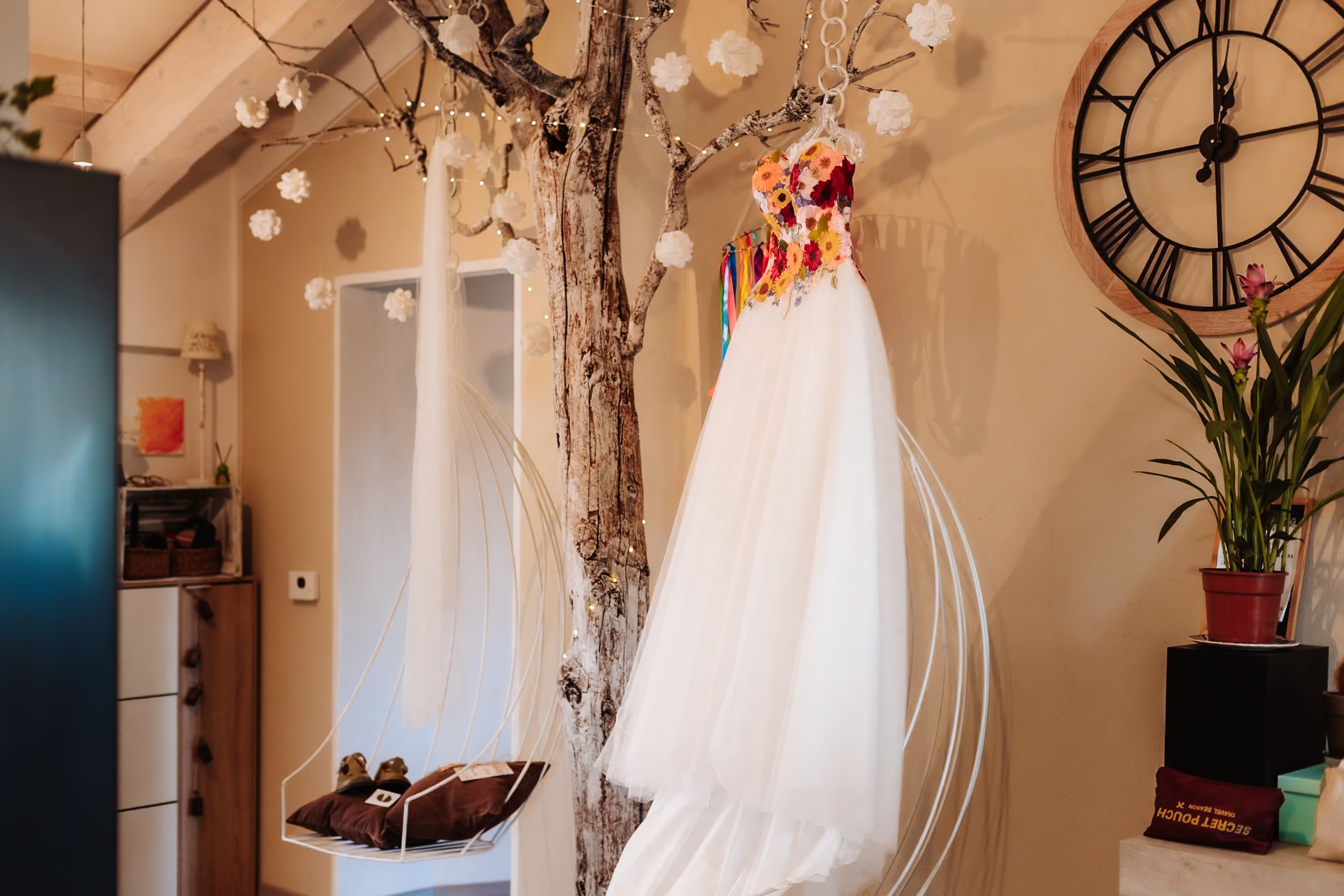 White wedding dress with colorful floral details hanging from a decorative tree branch indoors near a large wall clock.