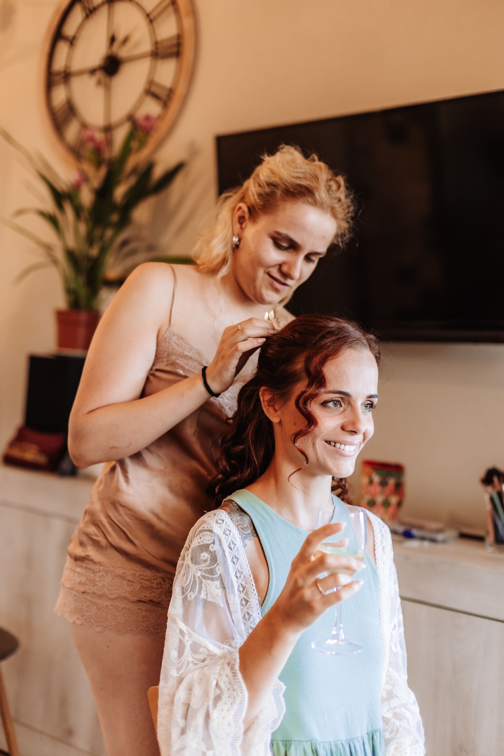 A woman styling another woman's hair while she smiles and holds a champagne glass during bridal preparations.