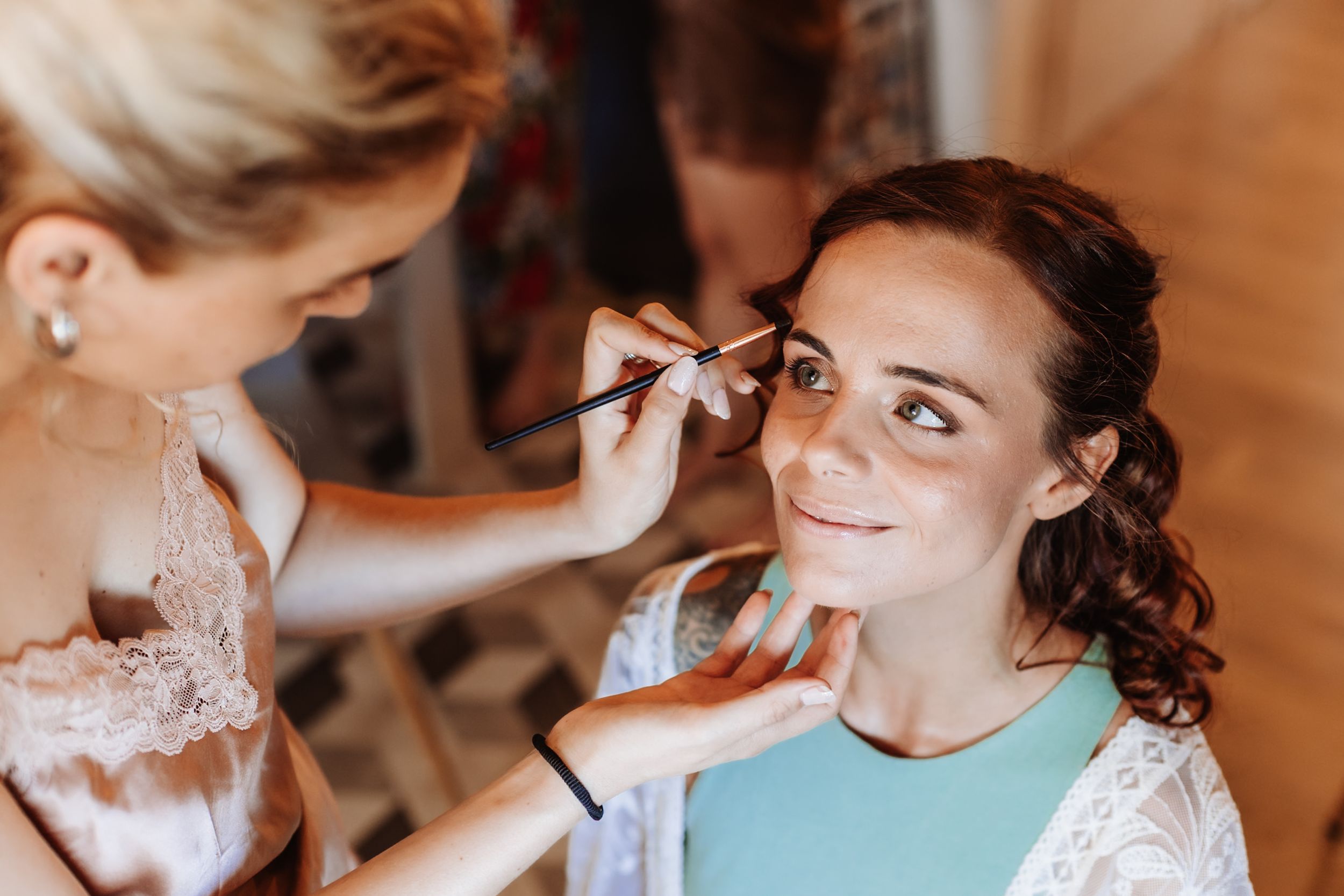 A makeup artist applying eyebrow makeup to a smiling bride during wedding preparations.