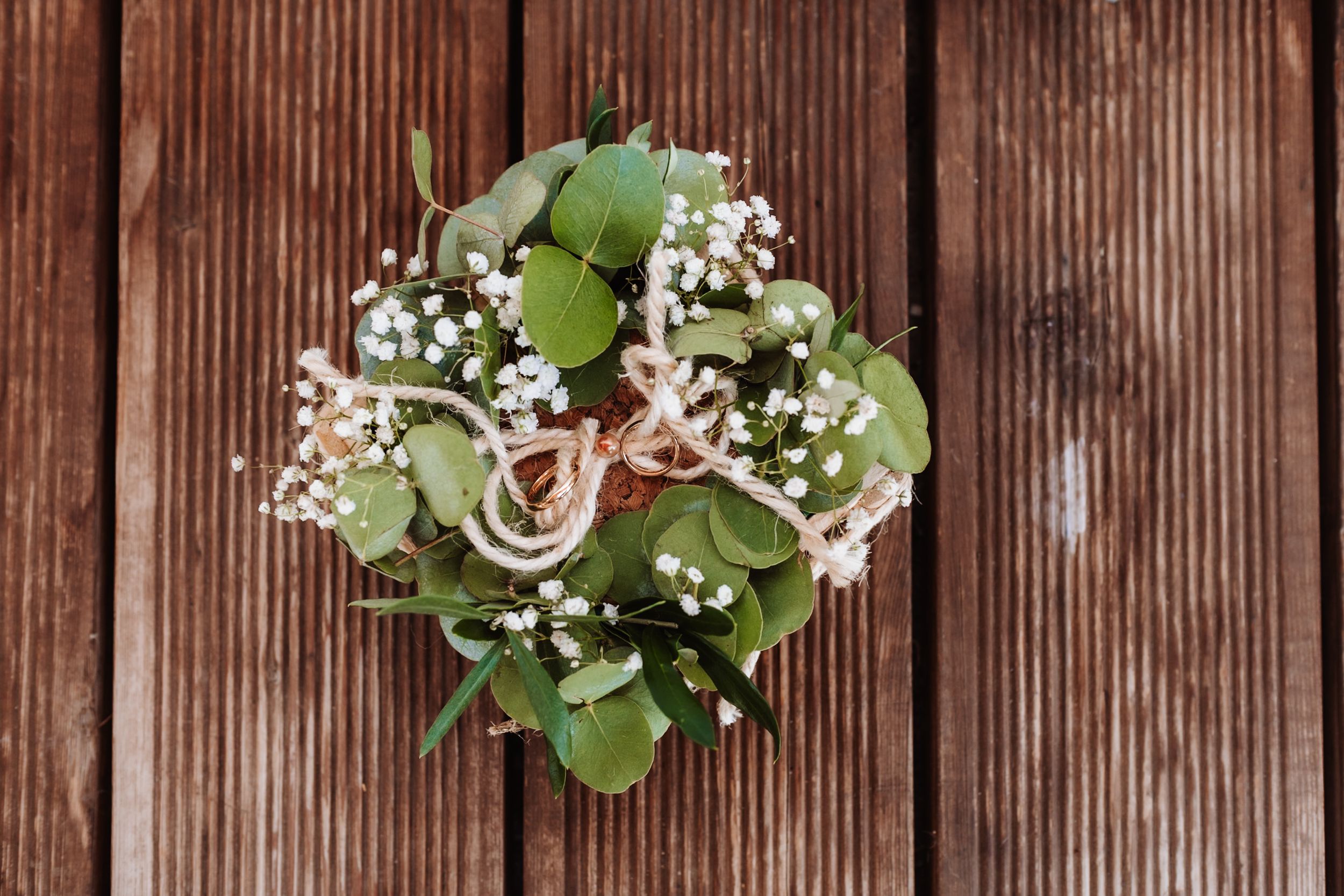 Two gold wedding rings tied with twine on a floral bouquet of eucalyptus and baby's breath on wood.