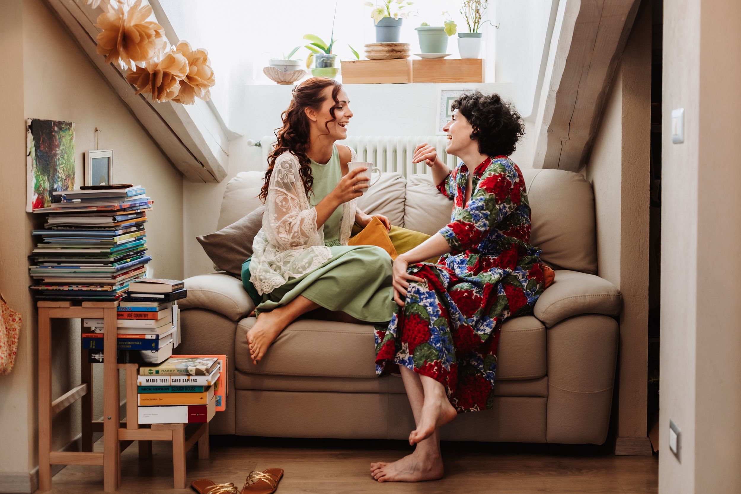 Two women sit on a couch chatting and laughing together in a cozy, book-filled living room.