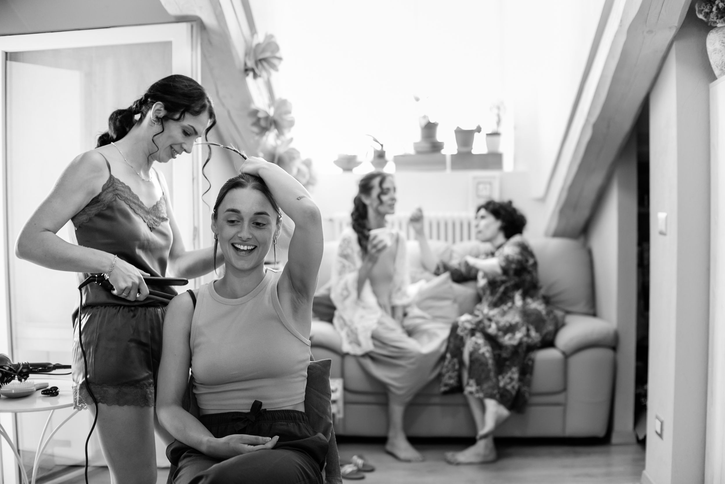 Black and white photo of women getting ready, one having hair styled while others chat on a sofa in a bright room.