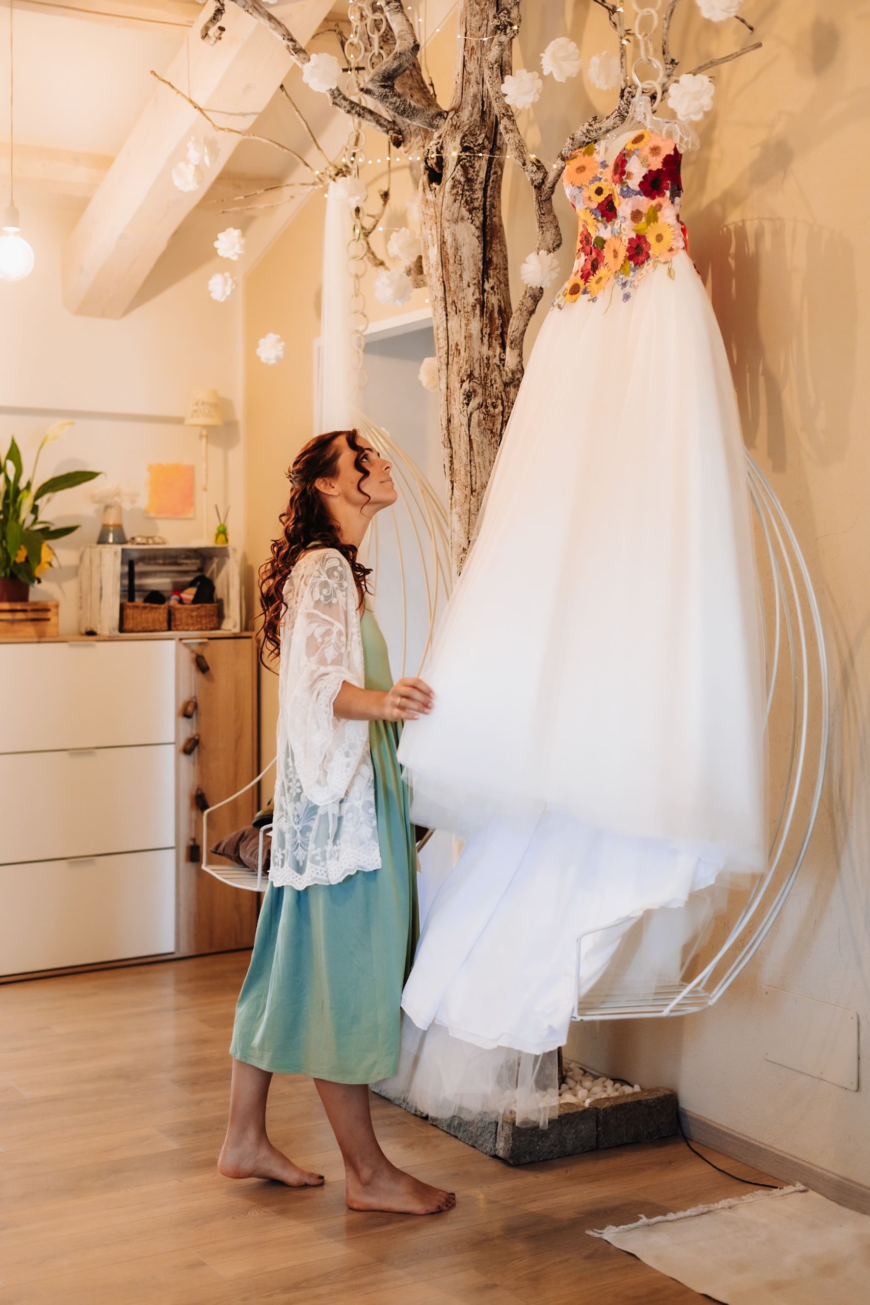 Barefoot bride admiring her floral wedding dress hanging from a decorative tree in a cozy bridal boutique.
