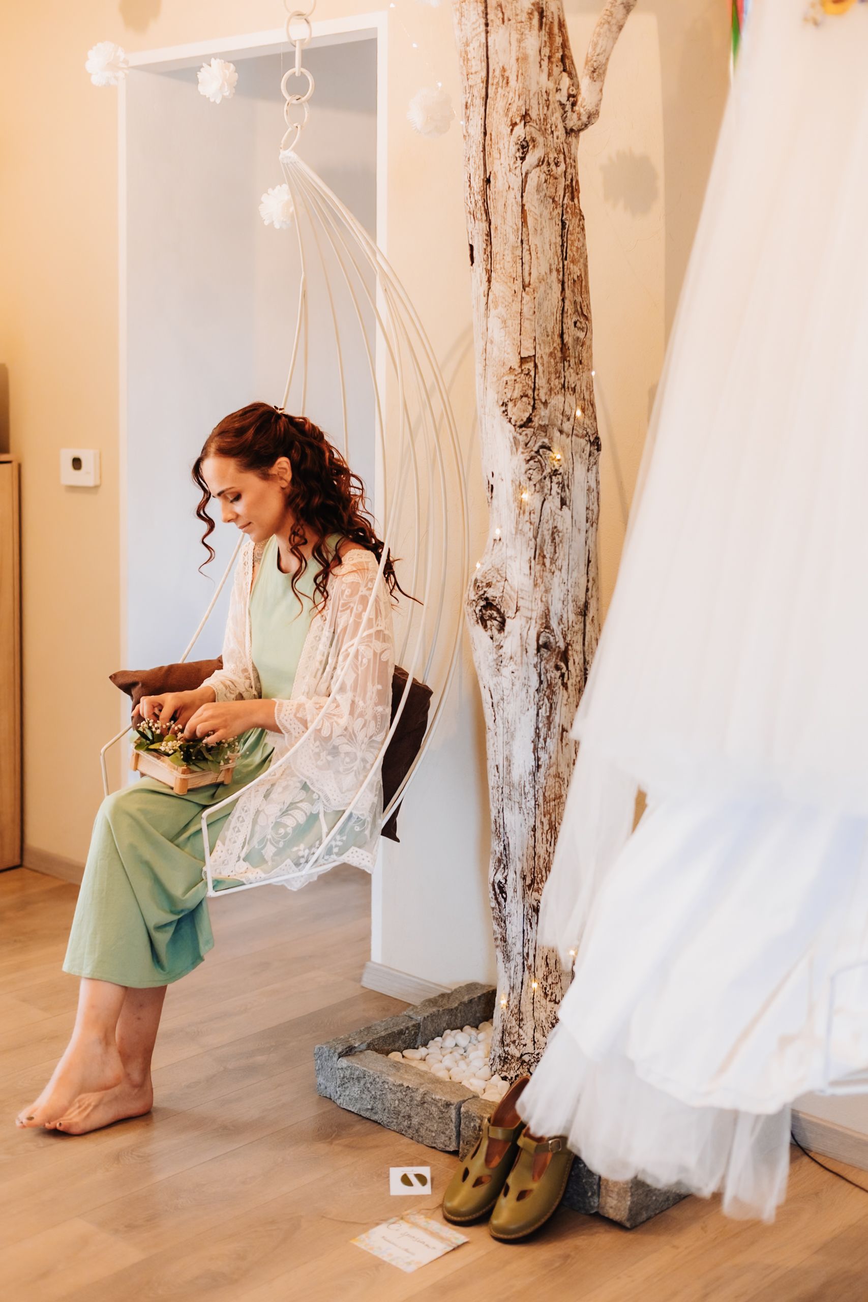 Woman in mint green outfit sits in macrame swing chair holding bouquet near rustic tree decor and wedding dress.