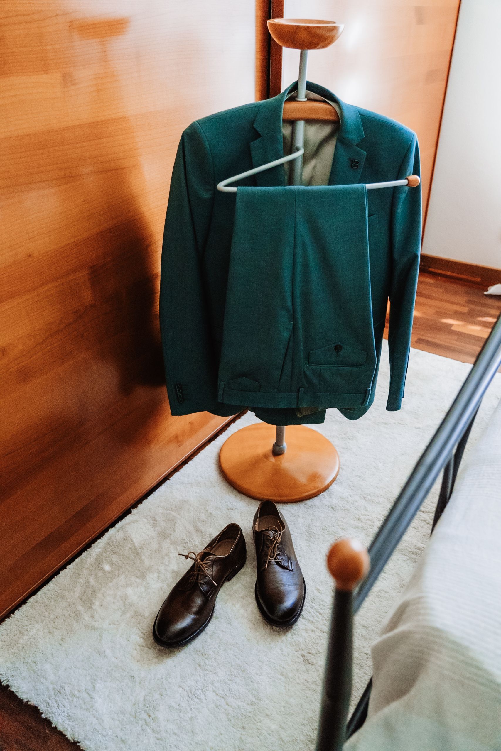 Green suit jacket and trousers on a valet stand with brown leather dress shoes on white rug.