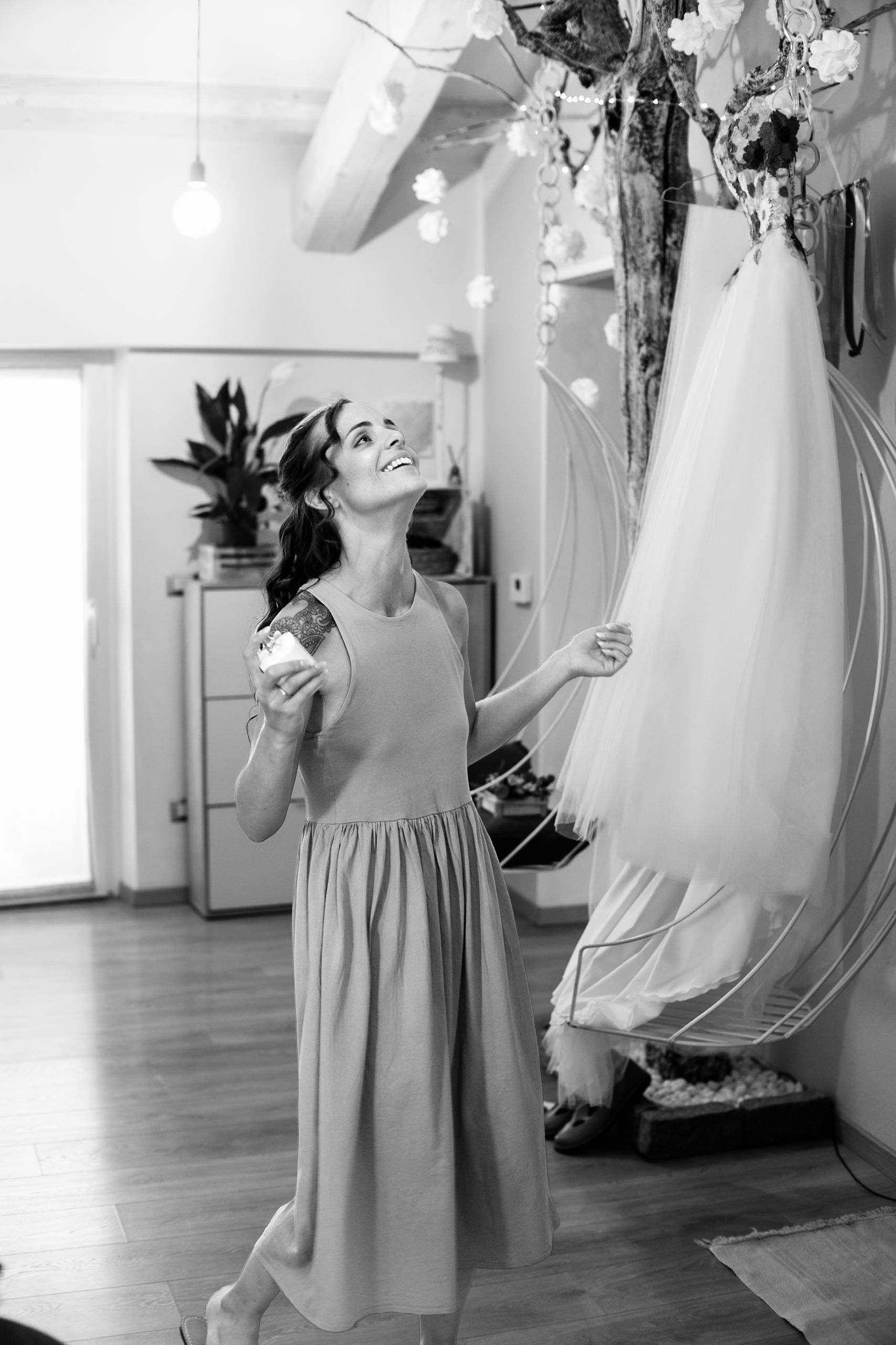 Black and white photo of a smiling bride in a long dress admiring her wedding gown hanging in a bridal boutique.
