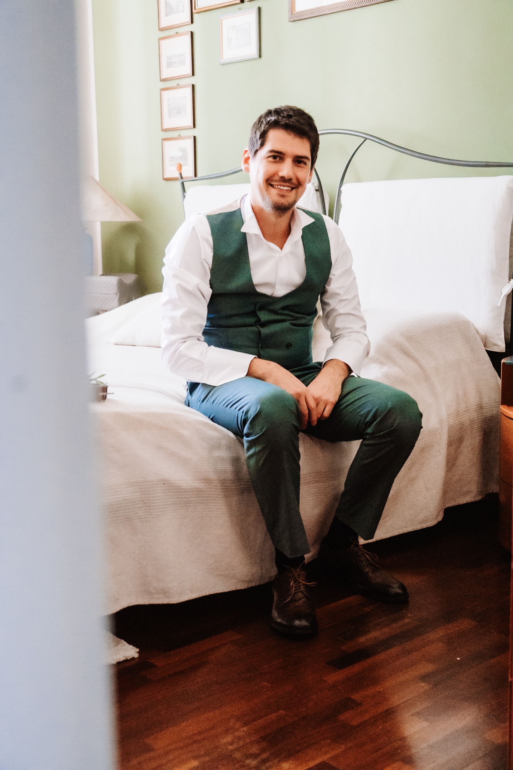 Smiling man in green vest and trousers sitting on a bed in a bright room with wooden floors.