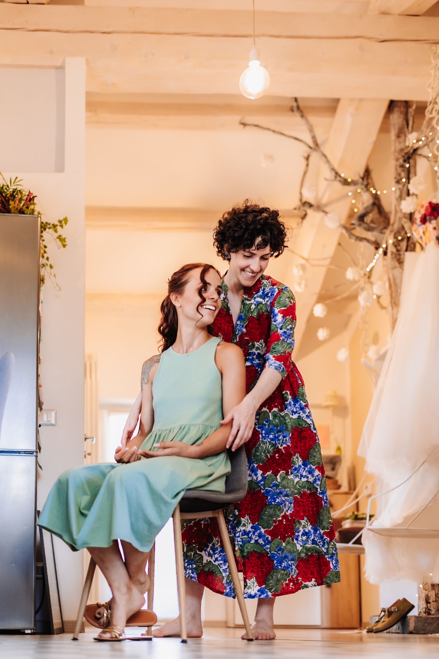 Two women in colorful dresses laughing together indoors, one seated on a chair in a warmly lit decorated room.