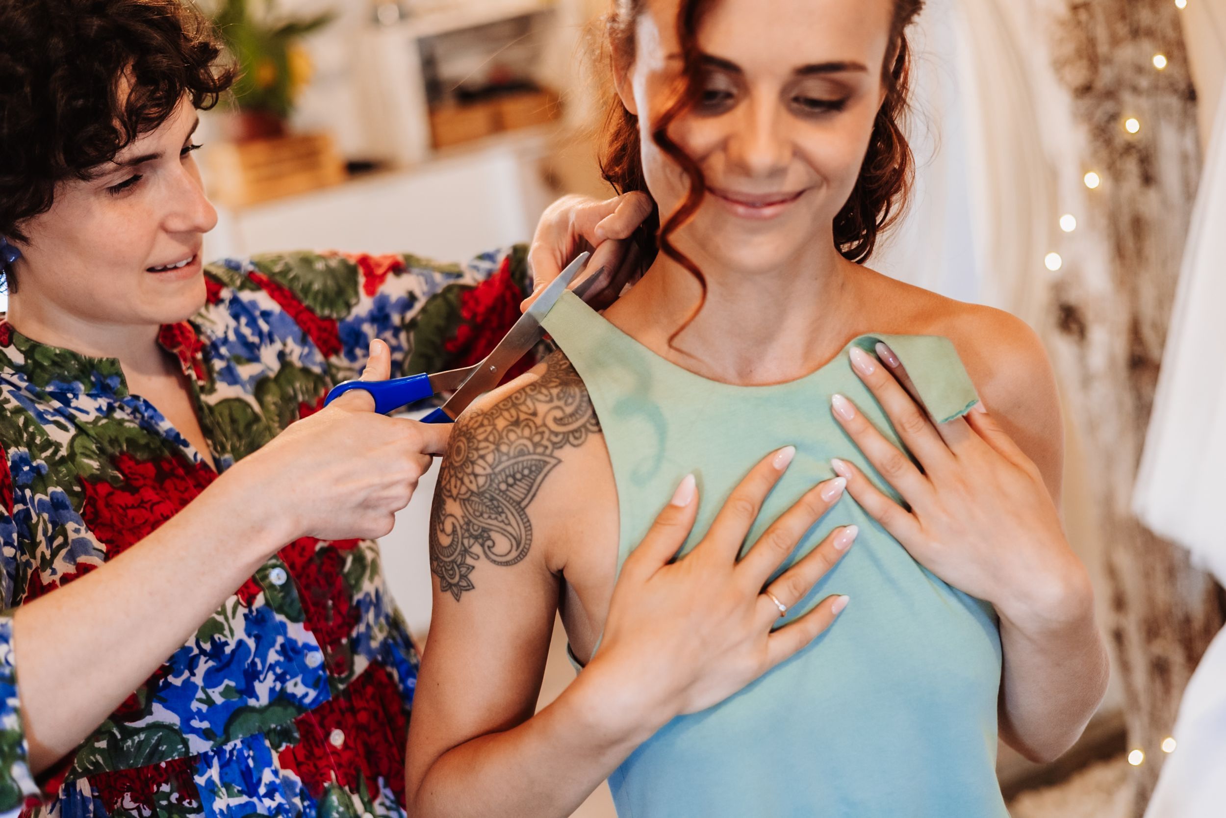 A seamstress adjusts a teal dress on a smiling woman with a tattoo in a bright bridal boutique.