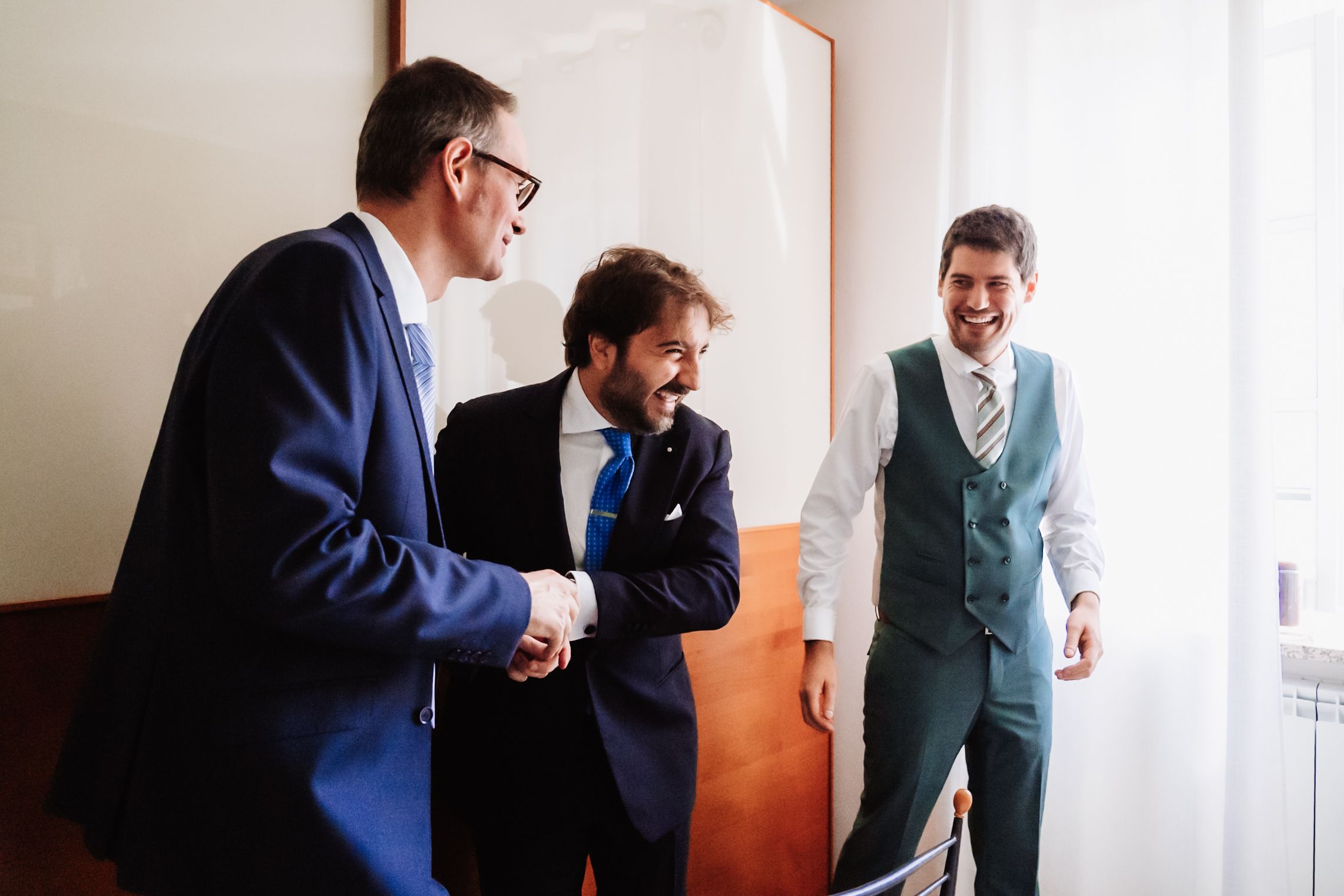 Three men in formal suits smiling and shaking hands in a bright room, likely at a wedding or business event.