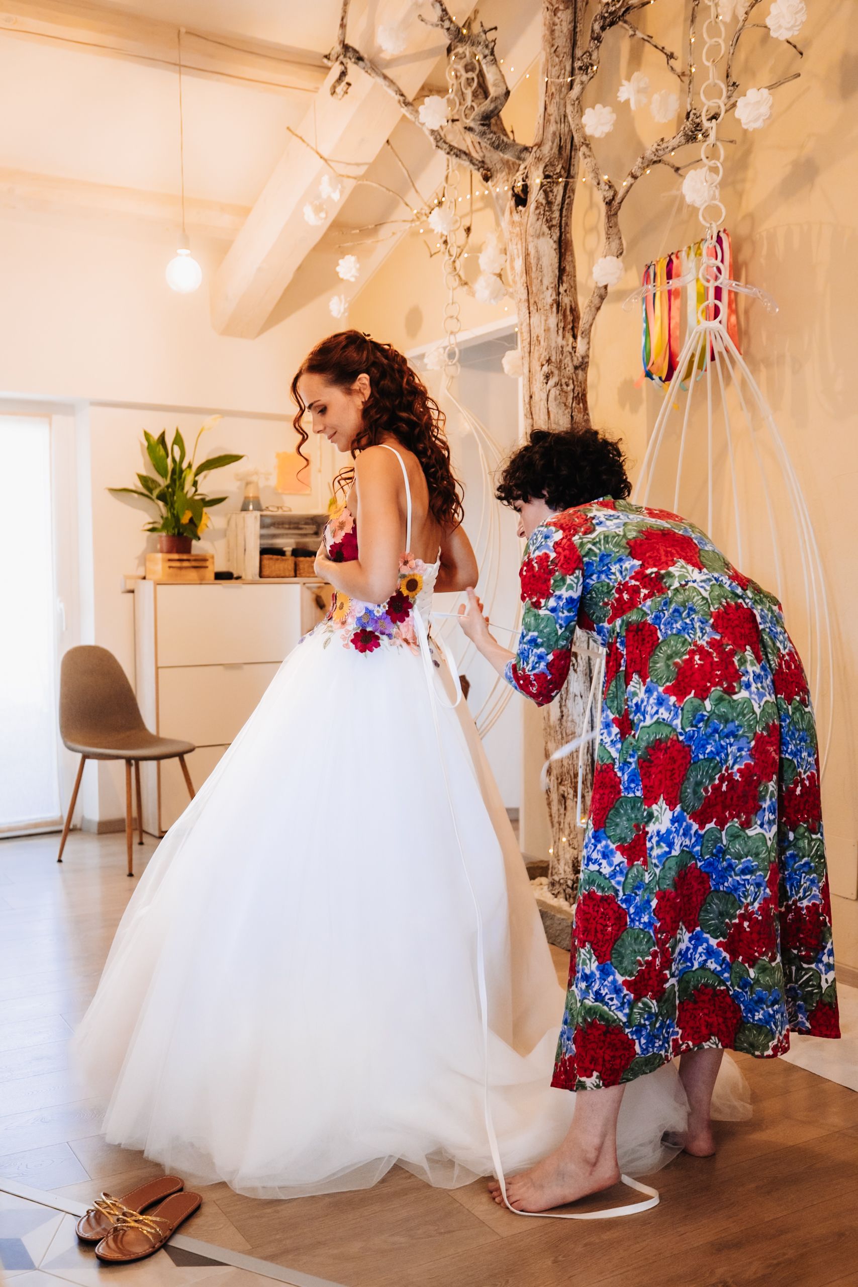 Bride in white ballgown with floral accents being helped by woman in floral dress inside decorated venue.
