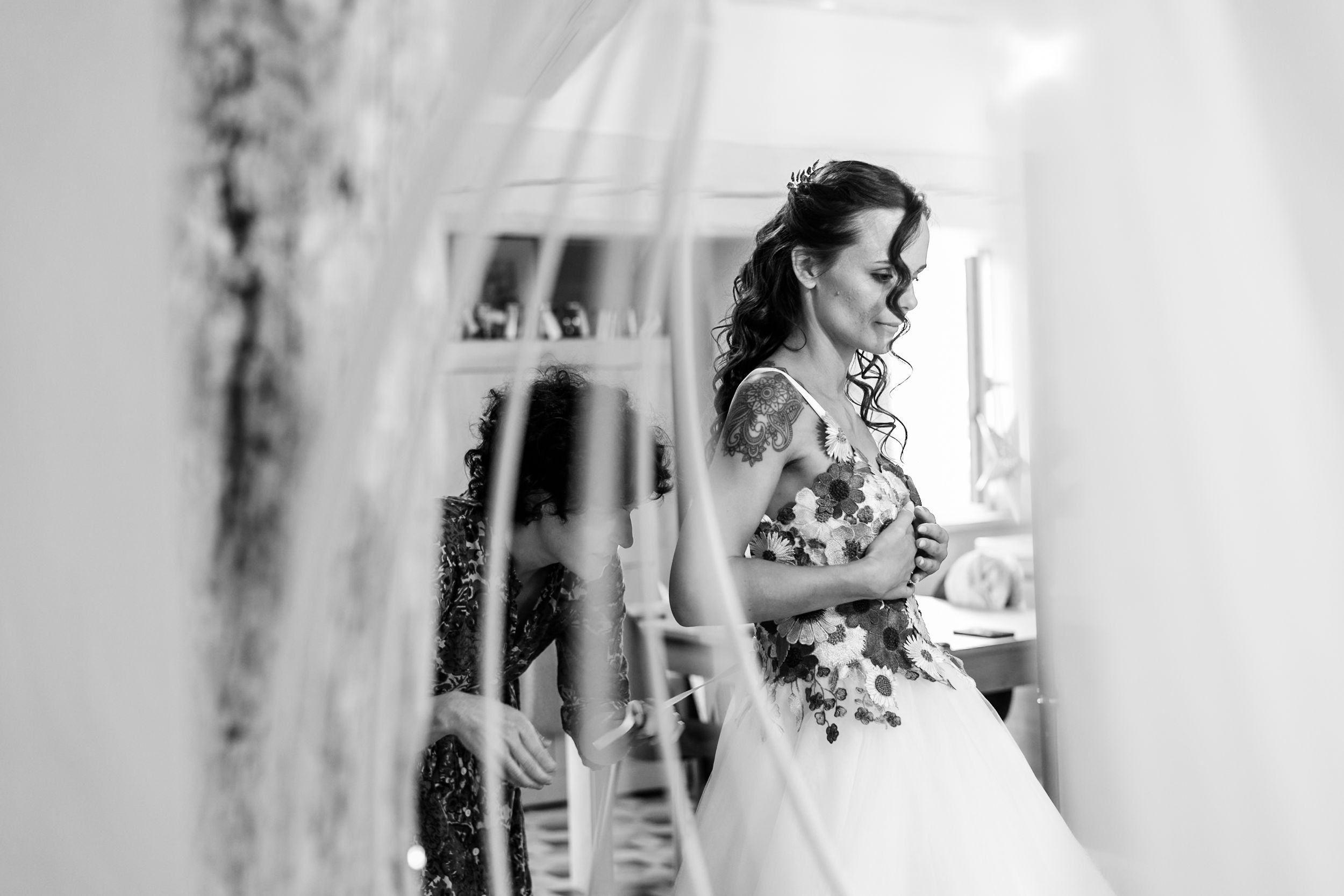 Black and white photo of a bride in a floral wedding dress holding a bouquet, with a helper adjusting her gown.