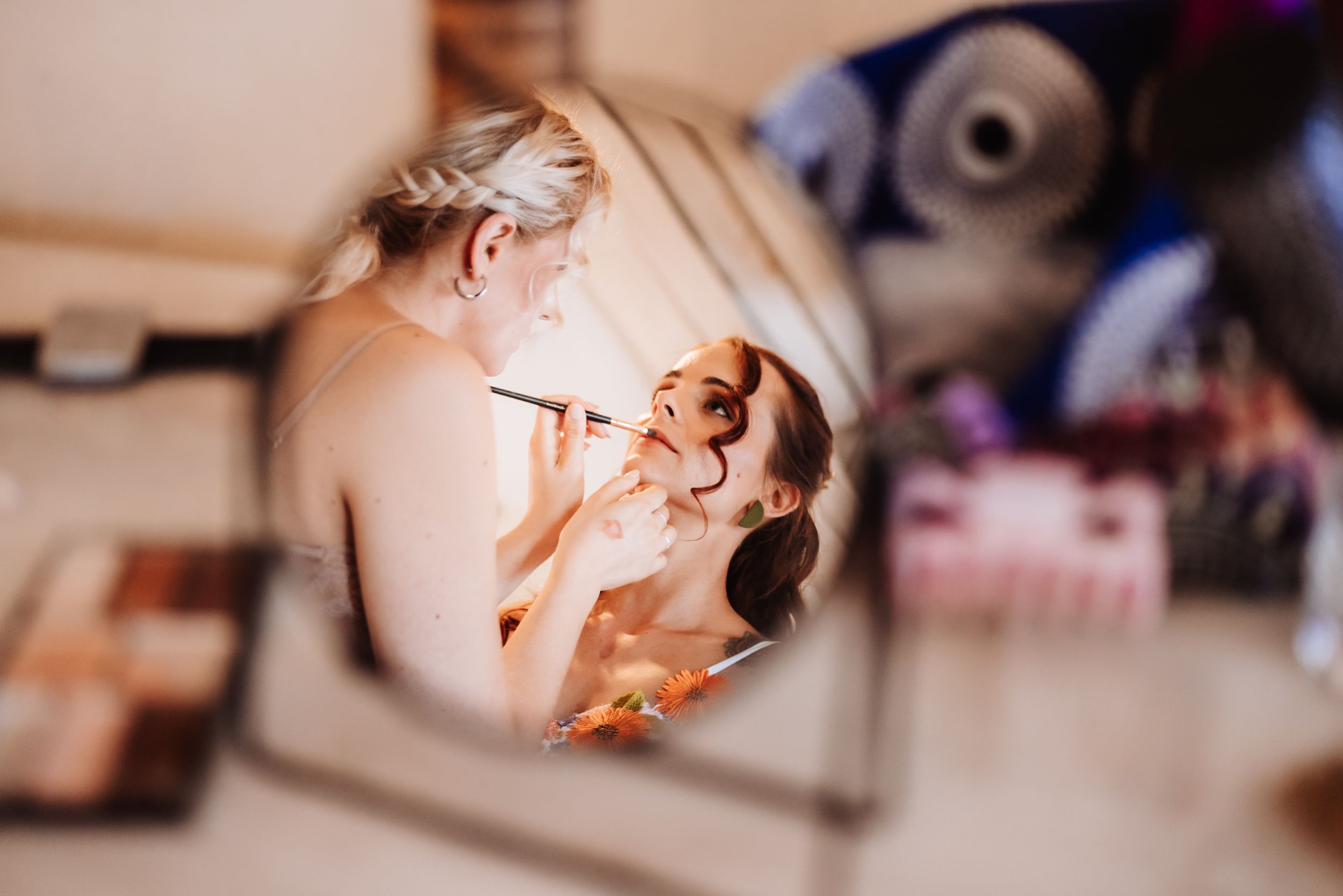 A makeup artist applying cosmetics to a bride, reflected in a round vanity mirror during wedding preparations.
