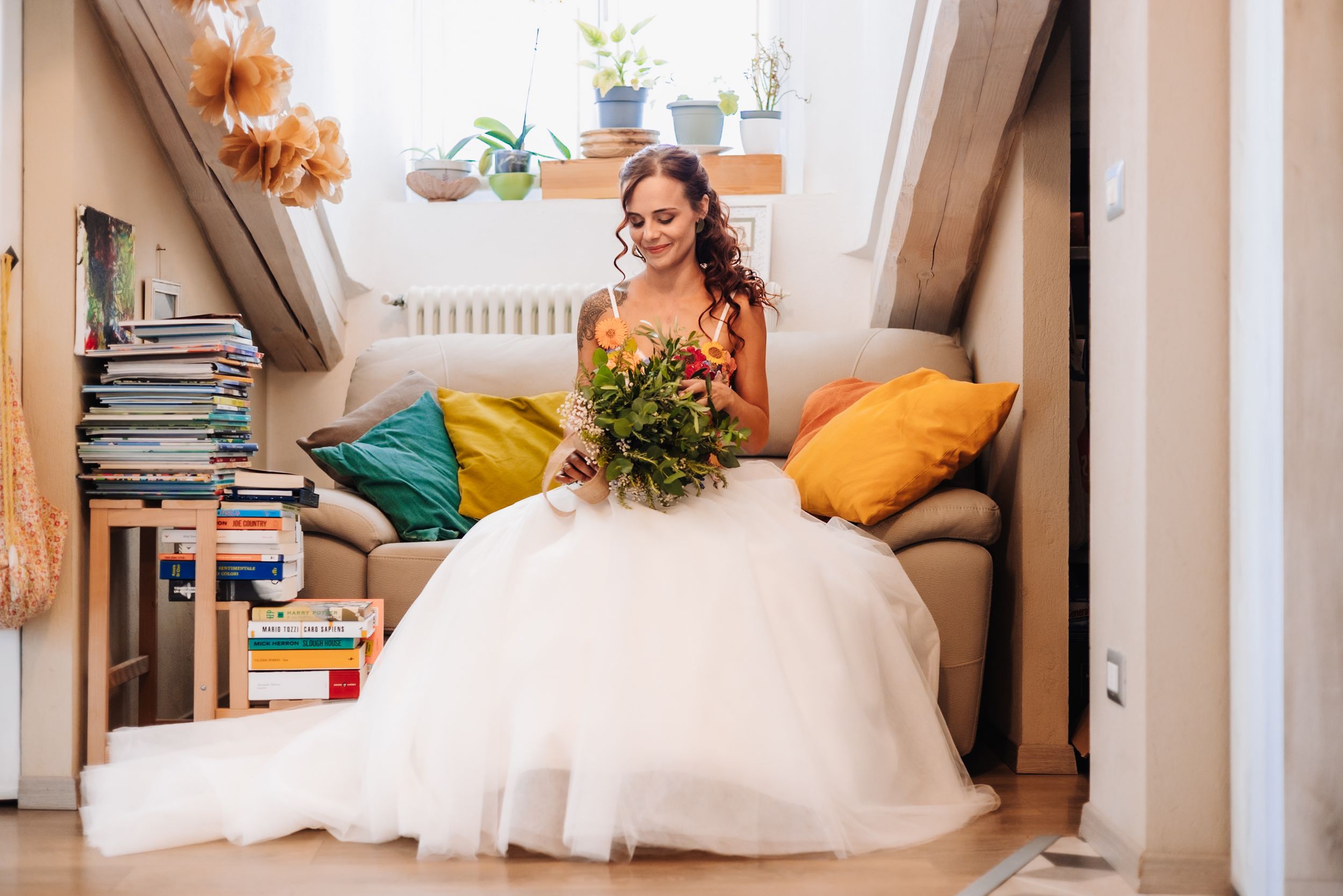Bride in white tulle gown sitting on sofa holding green bouquet in cozy room with colorful pillows.