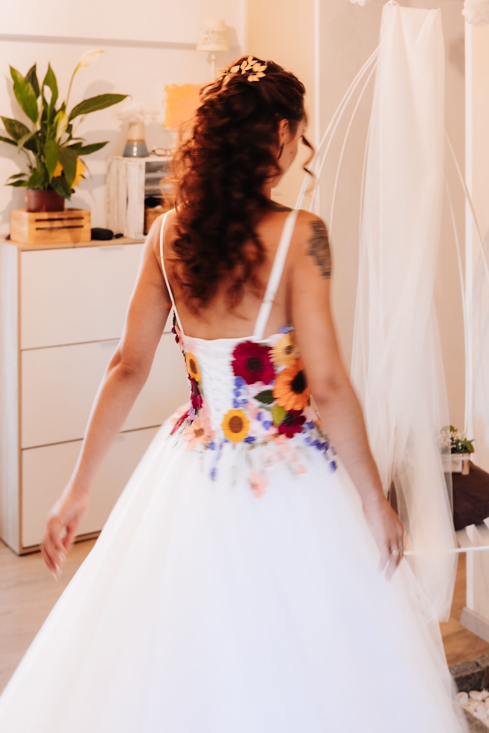 Bride in a white ballgown with colorful floral bodice and curly hair, viewed from behind in a bridal boutique.