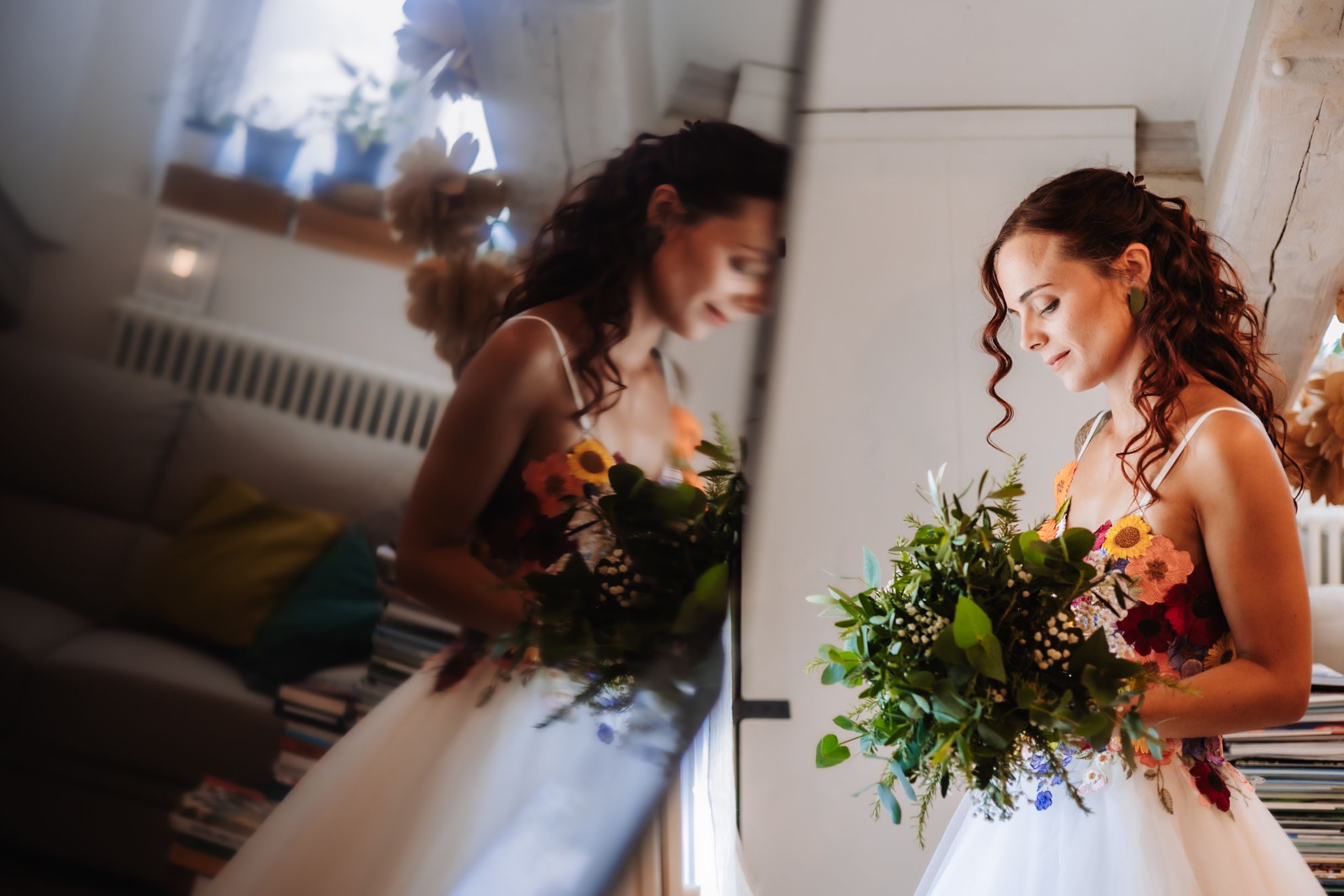 Bride in white gown holding colorful bouquet reflected in mirror before wedding ceremony.