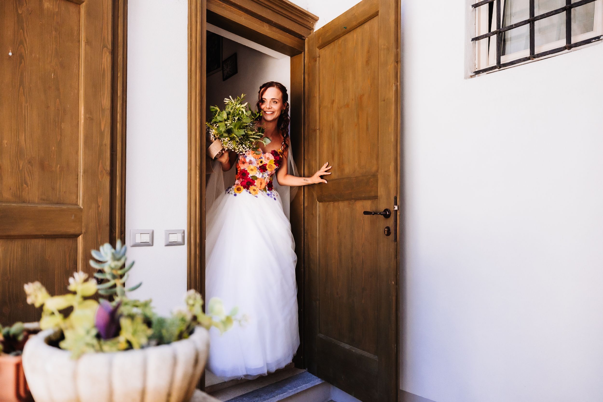 Bride in white tulle gown holding colorful floral bouquet standing in wooden doorway of rustic venue.