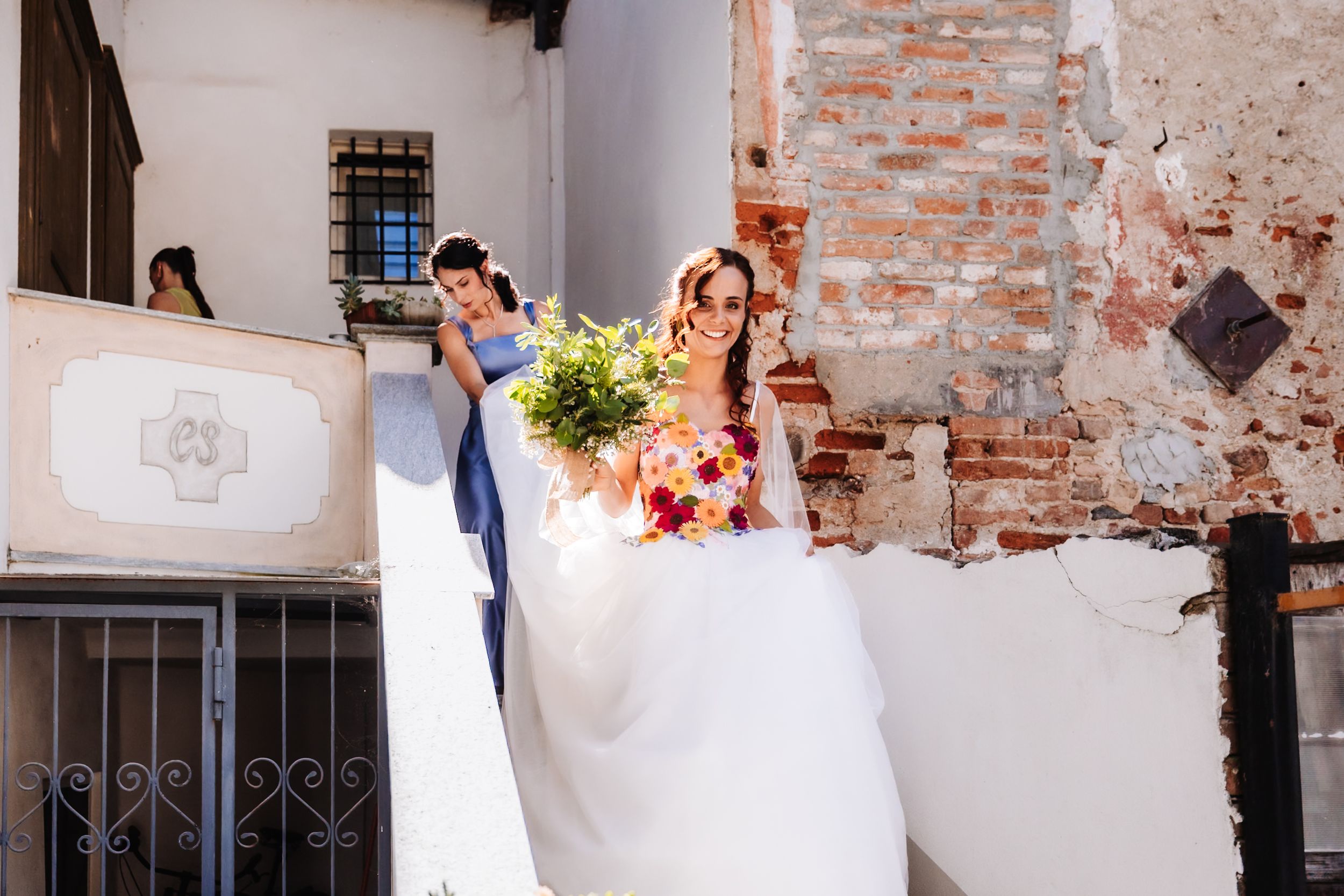 Bride in white gown holding colorful bouquet with groom on sunny outdoor staircase near rustic brick wall.