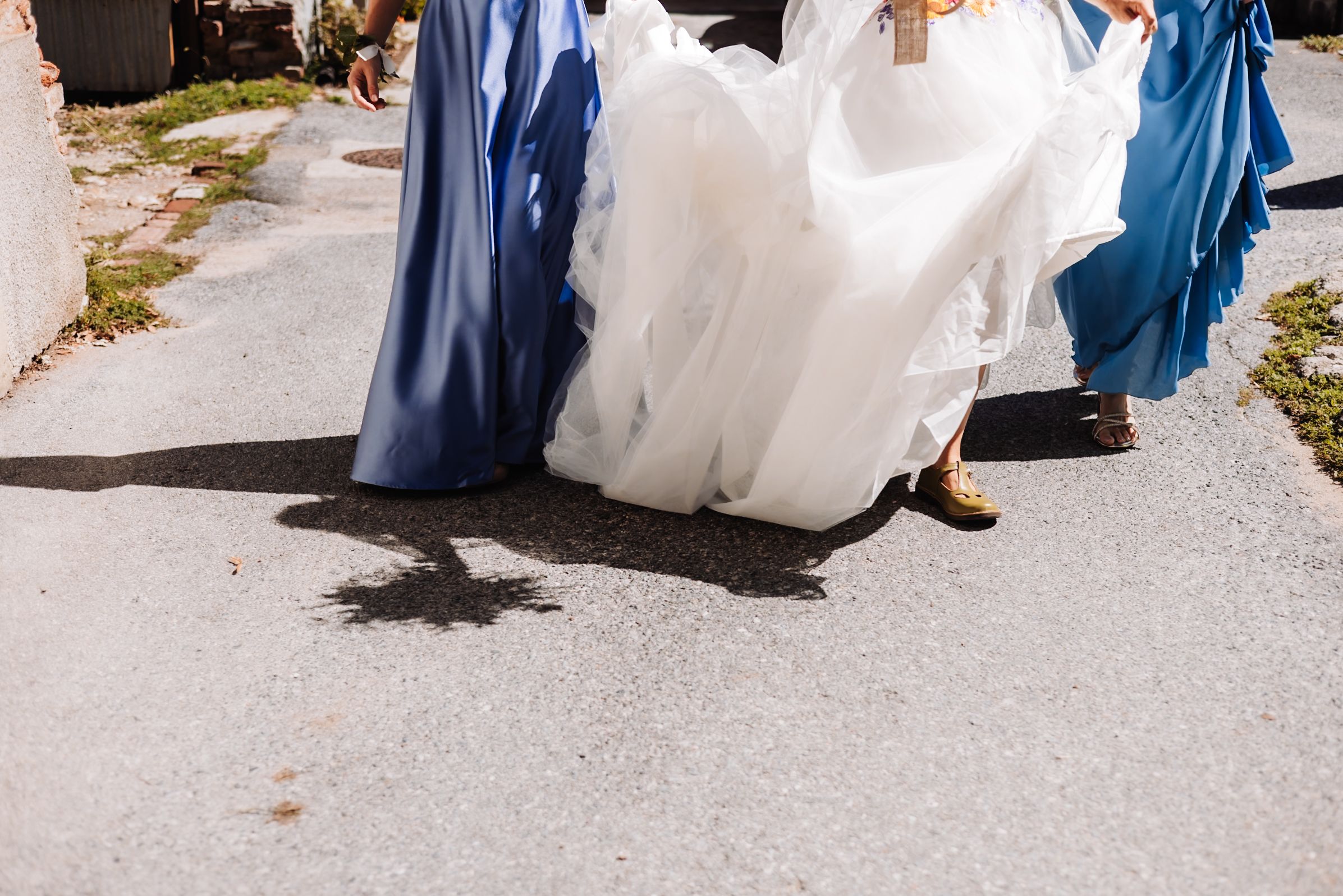 Bride in white gown walks with bridesmaids in blue dresses on sunny pavement, casting shadows below.