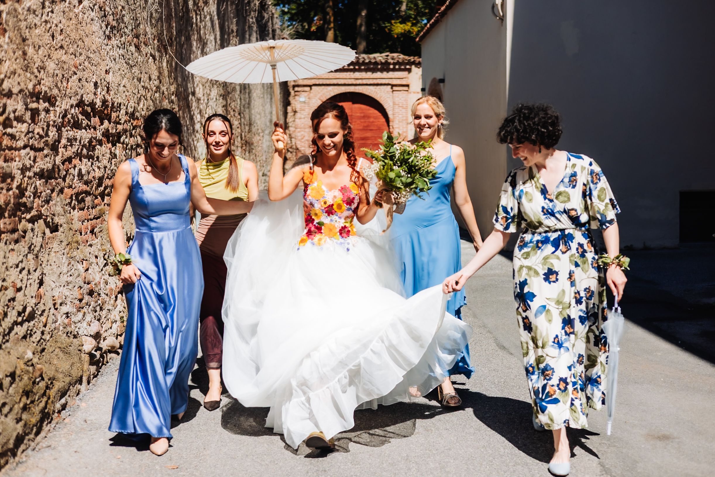 Bride in floral gown walks with bridesmaids in blue dresses, holding bouquet and parasol on sunny wedding day.