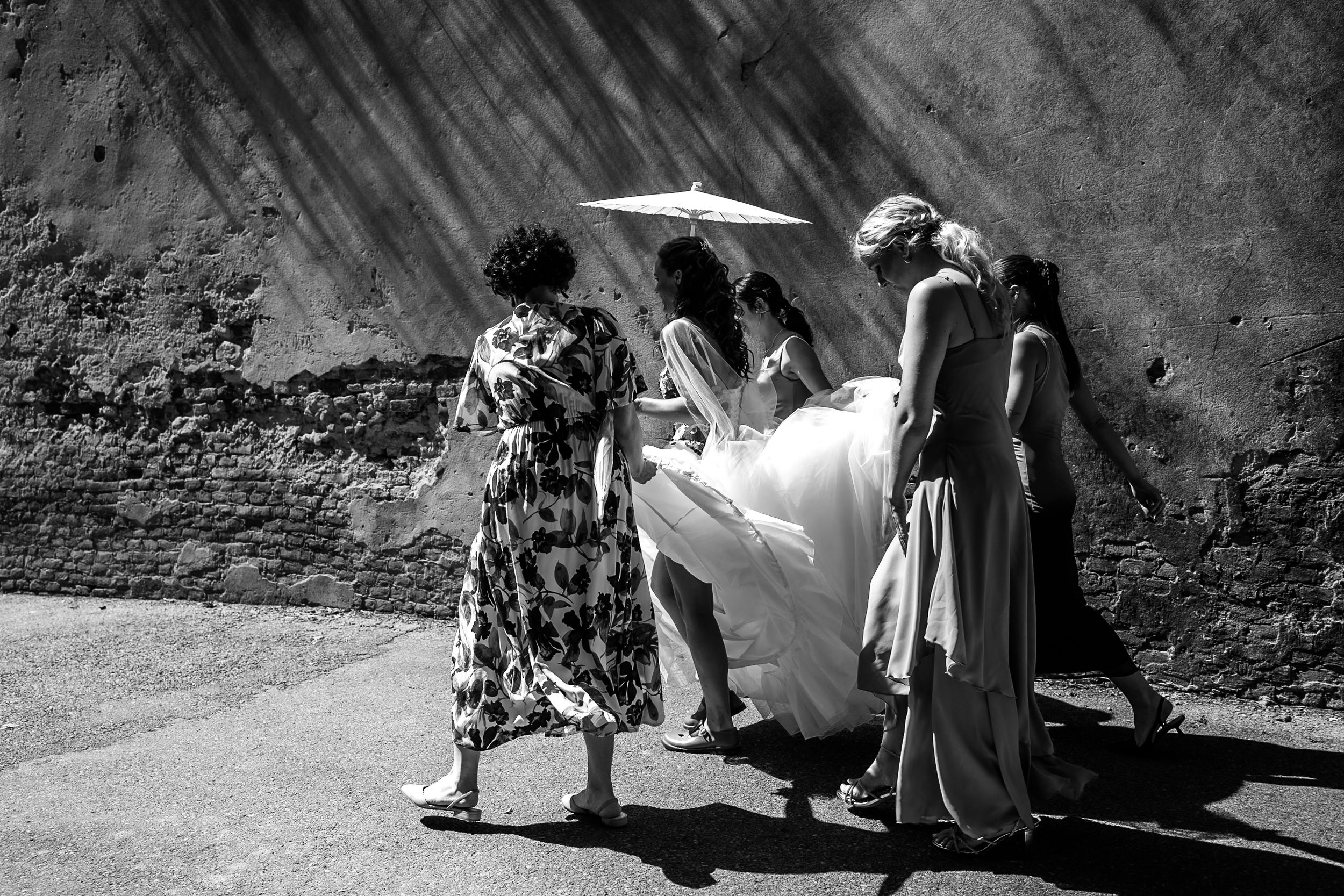 Black and white photo of a bride in a white gown walking with bridesmaids holding an umbrella outdoors.