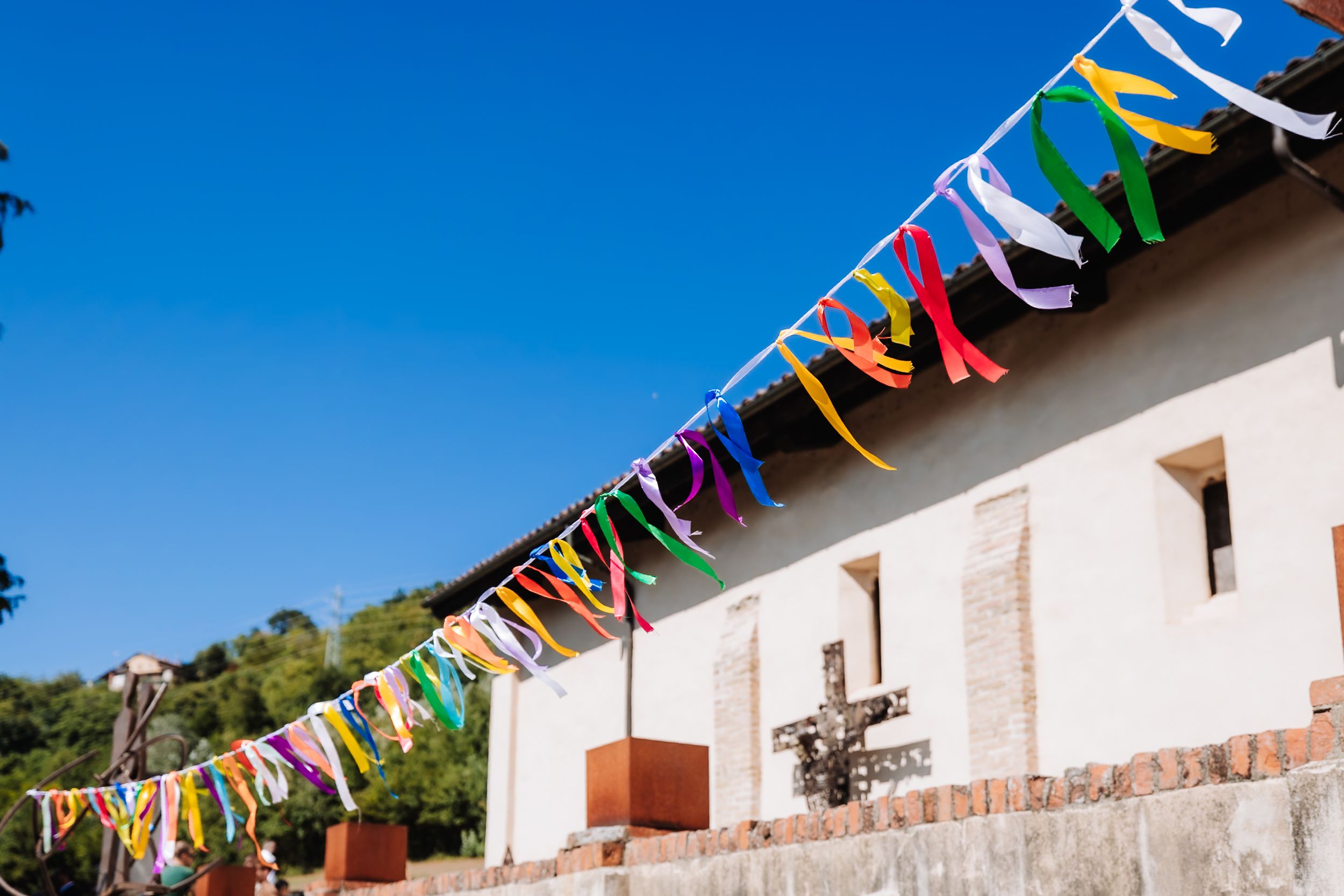 Colorful festive bunting flags strung along a white adobe church wall under a bright blue sky.