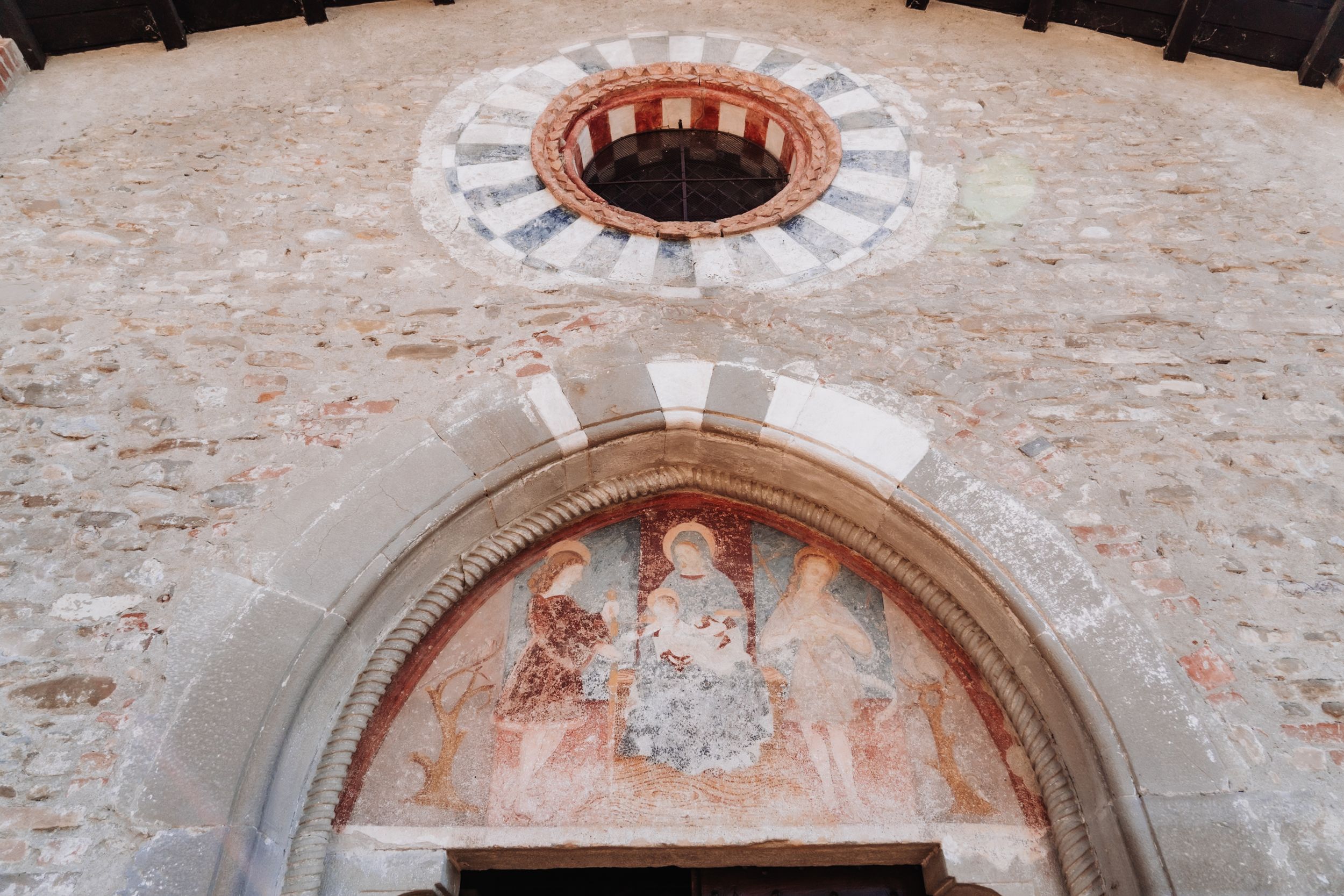 Ancient stone church facade with a rose window and faded fresco mural above an arched doorway.