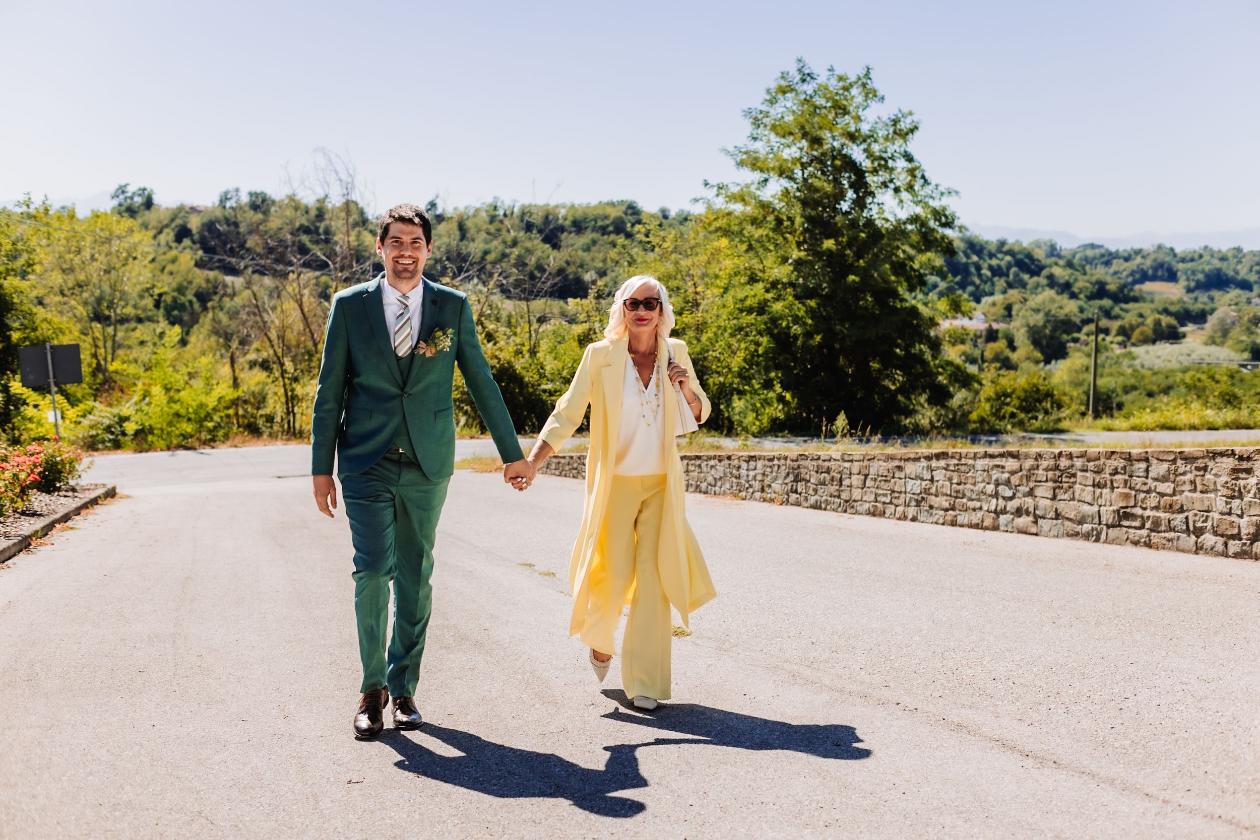 Couple in green suit and yellow dress walking hand in hand on a sunny road with scenic countryside views.